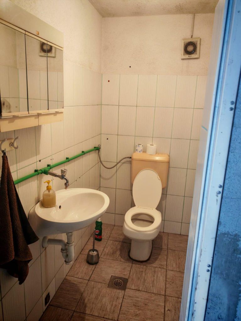 Small old bathroom with tiled walls, sink, toilet and visible green plumbing pipes before renovation.