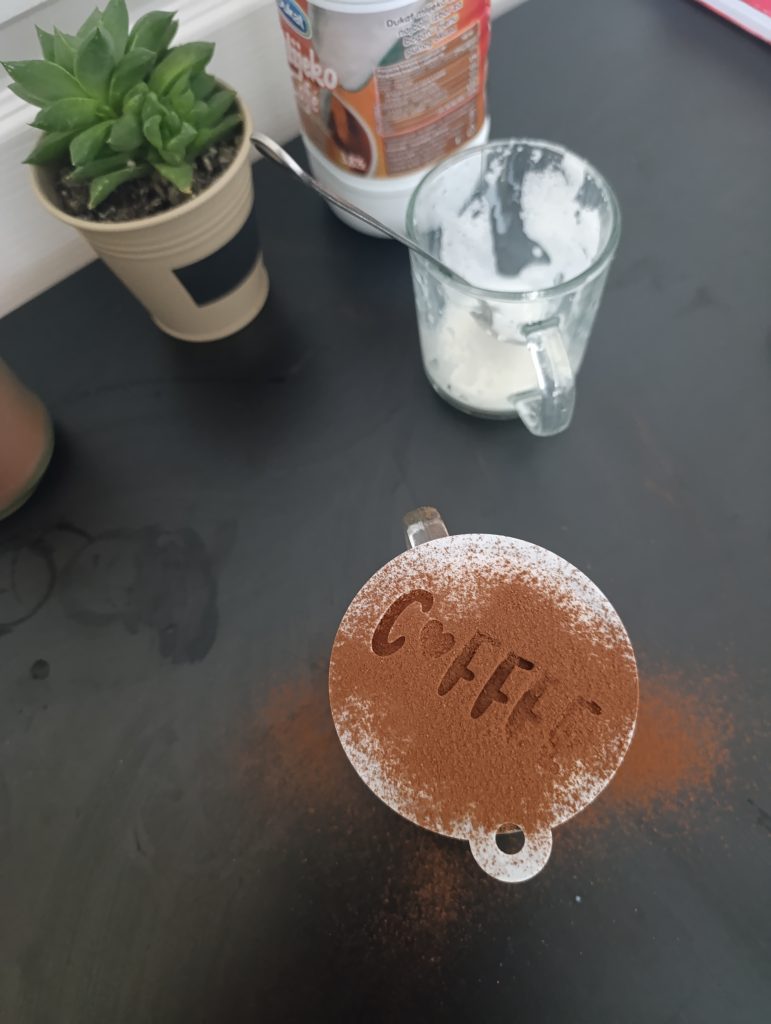 Dusting cocoa powder over milk foam through a coffee stencil to create a decorative design on top of a latte.