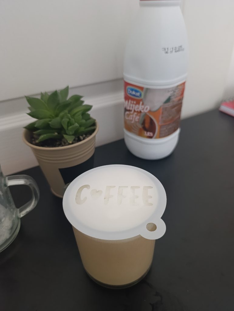 White coffee stencil placed on top of a glass mug filled with coffee, with a milk bottle and a small potted plant in the background.