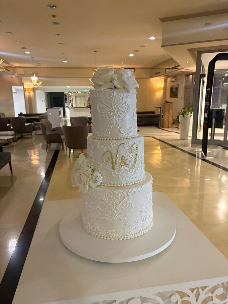 Three-tier white wedding cake decorated with an elegant Damask Stencil, gold initials and white flowers