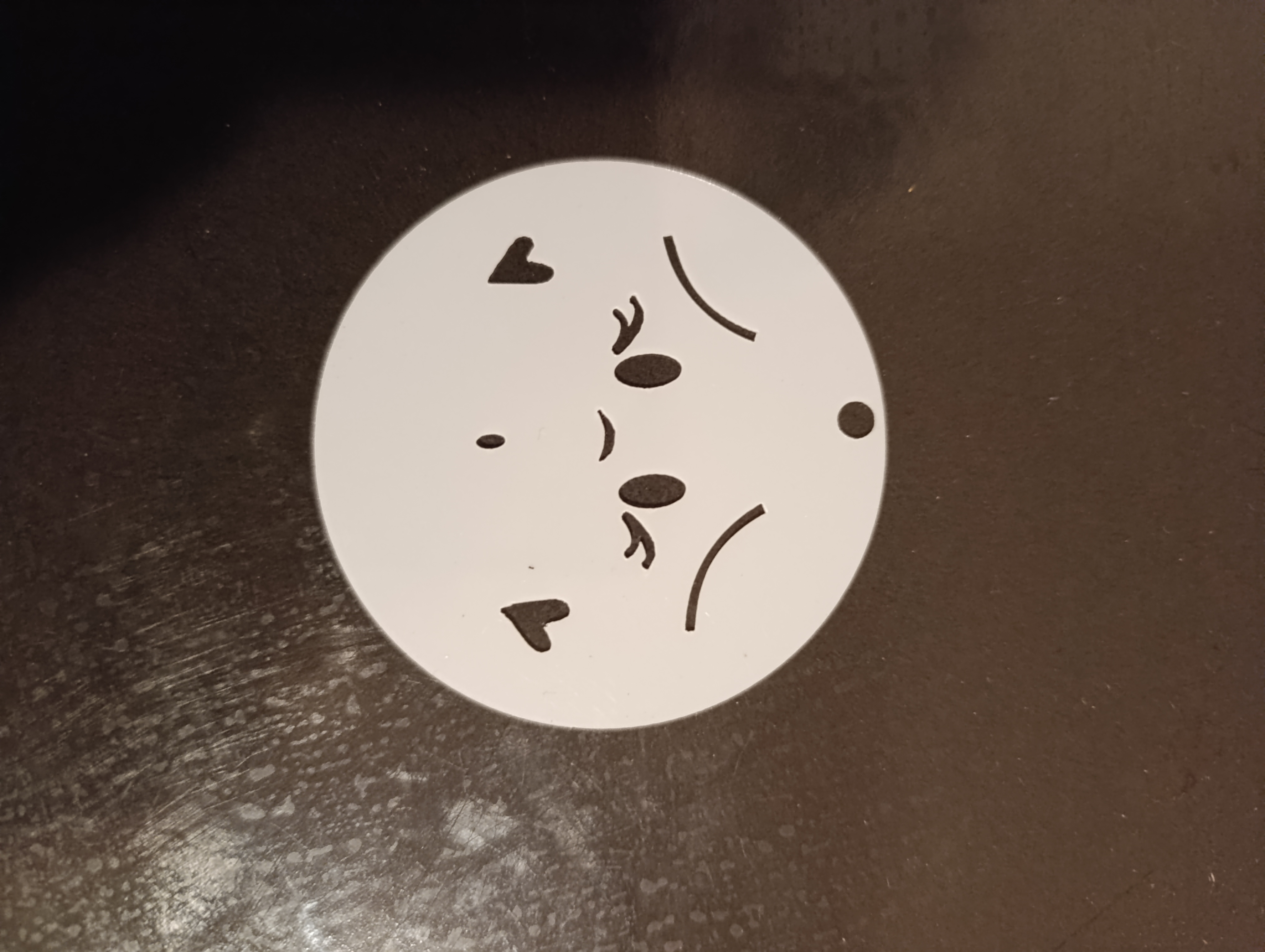 Circular white stencil featuring a shy facial expression with small heart shapes on the cheeks and a hanging hole.