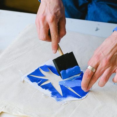 Person applying blue paint with a foam brush over a stencil on a canvas tote bag.