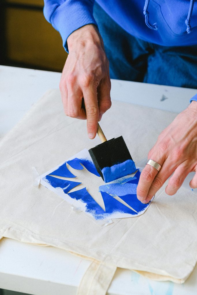 Person applying blue paint with a foam brush over a stencil on a canvas tote bag.
