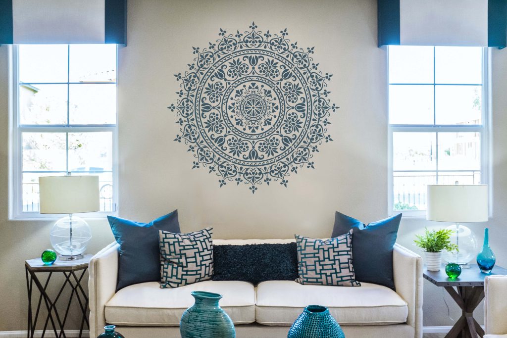 Living room wall decorated with a large circular mandala stencil design centered above a white sofa between two windows.