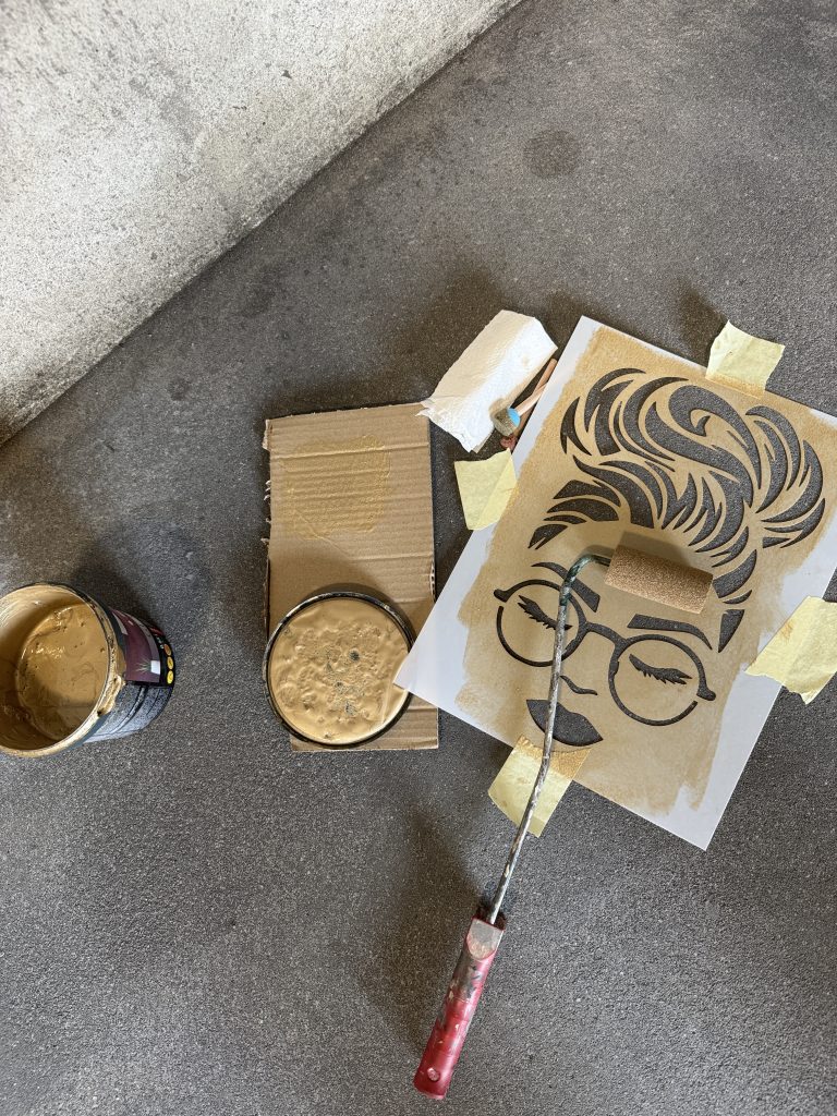 Top view of a portrait stencil taped to a concrete surface while beige paint is applied with a roller, with an open paint can nearby.