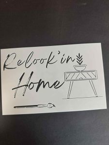 Stencil template with the phrase “Relookin’ Home” and a simple furniture illustration, displayed on a dark surface.