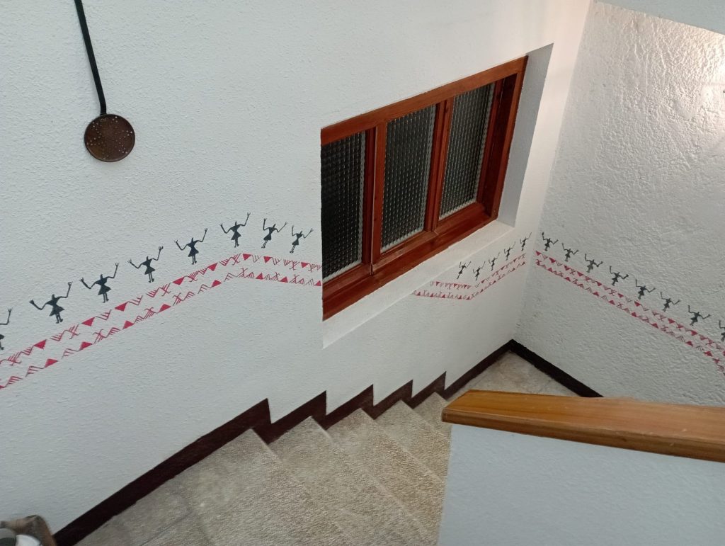 Staircase interior decorated with a continuous stencil border of small human figures and red geometric motifs running along the white walls near a wooden-framed window.