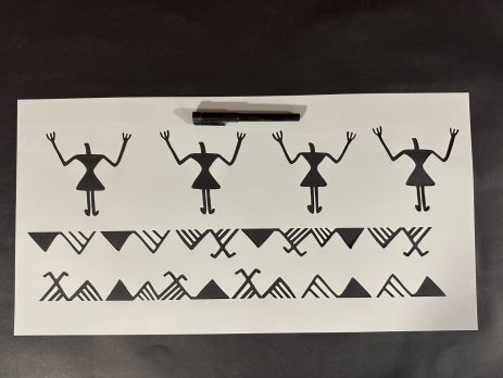 Stencil sheet featuring repeating tribal-style human figures and geometric border patterns placed on a table with a pen above it.